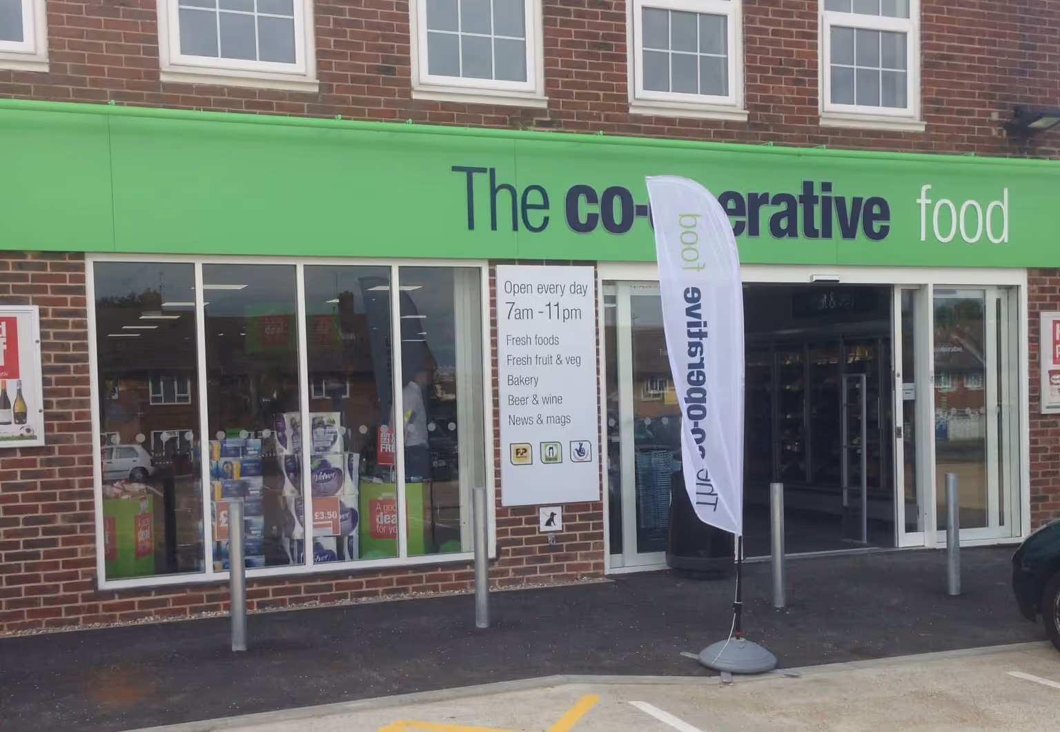 The Co-operative food store storefront with green signage, windows, and opening hours displayed on brick building.