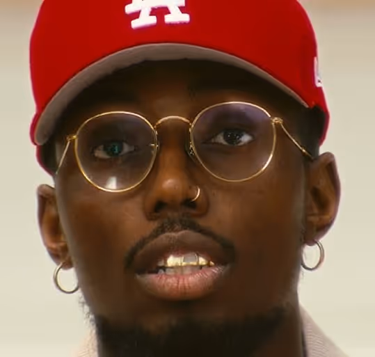 Close-up of a man wearing a red cap, round glasses, gold grills, and hoop earrings.