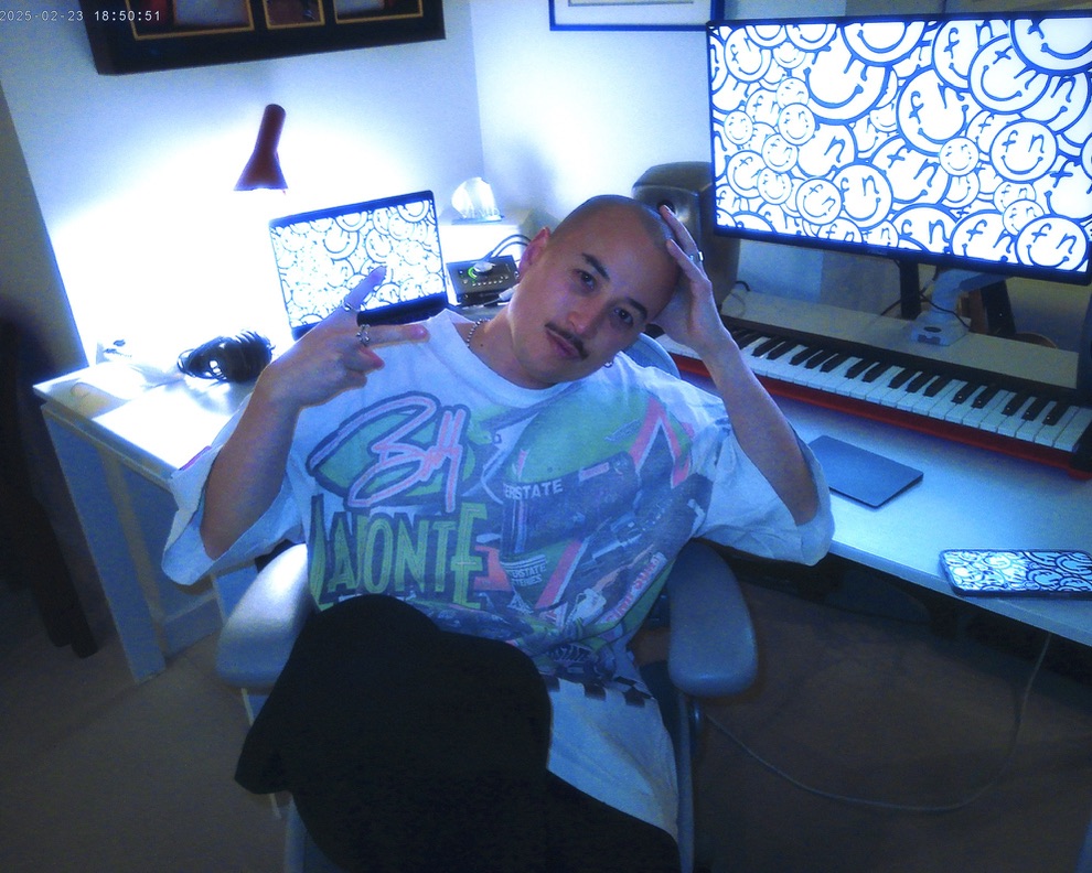 Person with a mustache and shaved head making a peace sign while sitting in front of a keyboard and two screens displaying smiley face patterns.