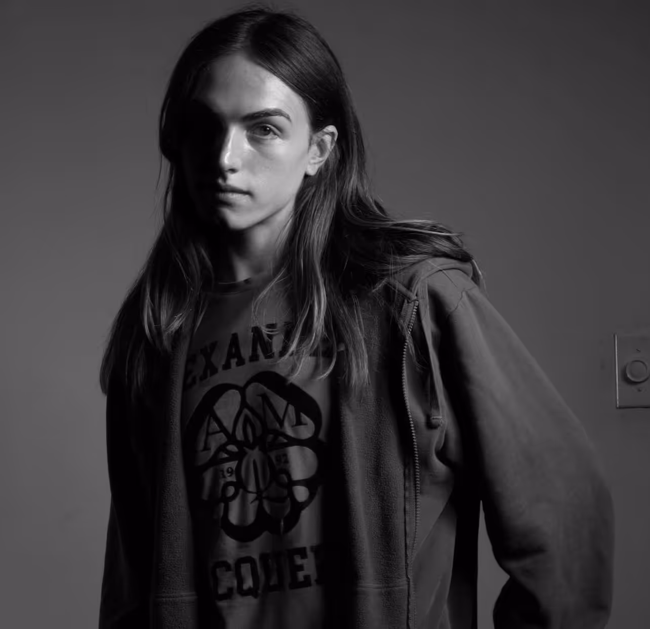 Black and white portrait of a young person with long hair wearing a zip-up hoodie and a graphic t-shirt, lit from the side against a plain background.