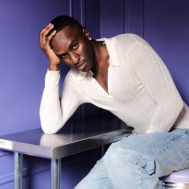 Man with short hair and earring leaning on a metal table wearing a white long-sleeve shirt and light blue jeans against a purple wall.