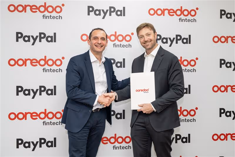 Two men in business suits smiling and shaking hands in front of a backdrop featuring Ooredoo fintech and PayPal logos, one holding a folder with the Ooredoo fintech logo.