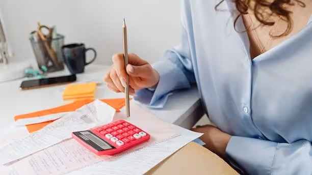 Woman holding a pencil sits at a desk with receipts, papers, and a pink calculator.