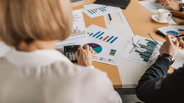 People review printed charts and graphs at a table, with a calculator and coffee cup nearby.