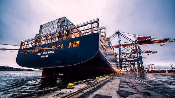 Large container ship docked at a port, with cranes and cargo containers in the background.
