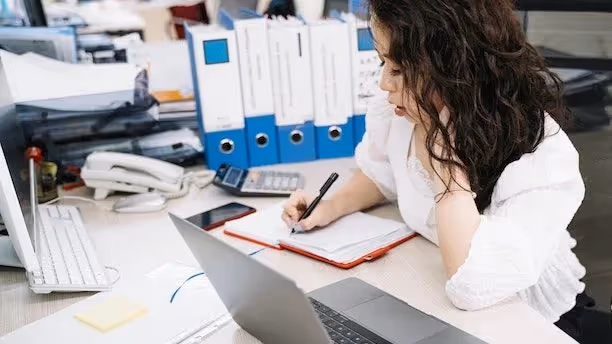 Woman at a desk writing in a notebook with a laptop, phone, and binders nearby.