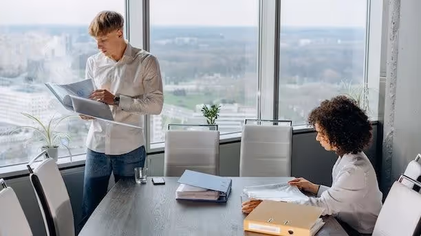  A male and female coworker stand near a conference table with documents, looking over paperwork.
