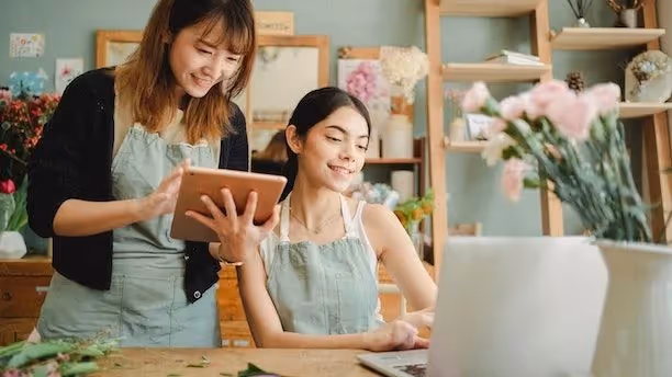 Two women in a flower shop work together using a tablet and laptop.