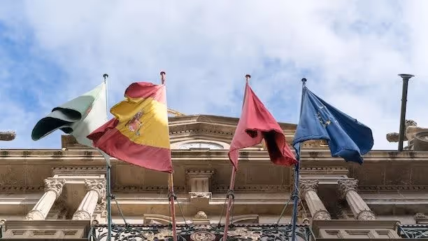 Flags of Spain, the EU, and regional banners flying in front of a historic building.