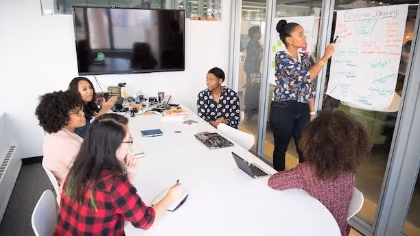 A woman leads a meeting, pointing to a flipchart as five colleagues listen and take notes.