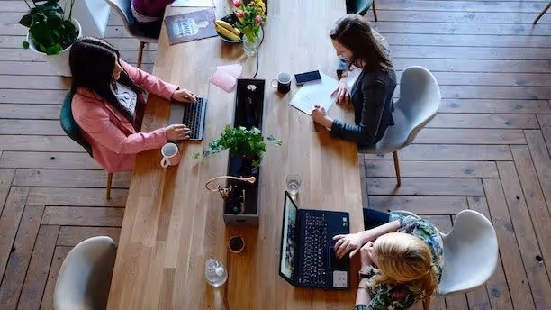 Three women work at a long wooden table with laptops, notebooks, and coffee in a shared office space.