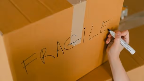 A person writes the word 'fragile' on a brown cardboard box.