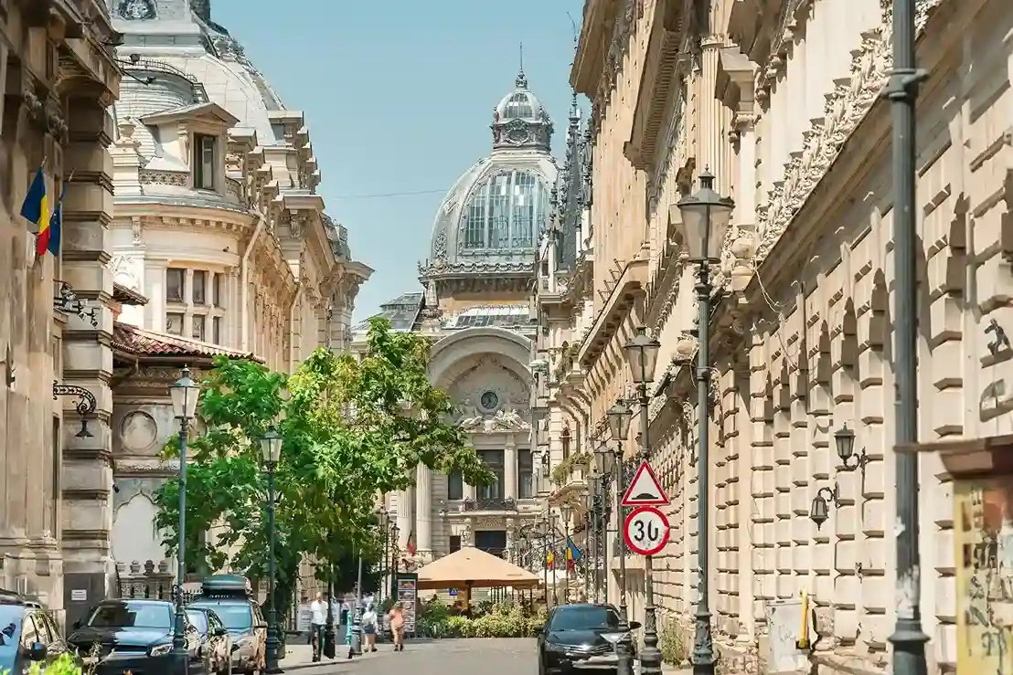 Historic street in Bucharest with ornate buildings, dome, and Romanian flags.