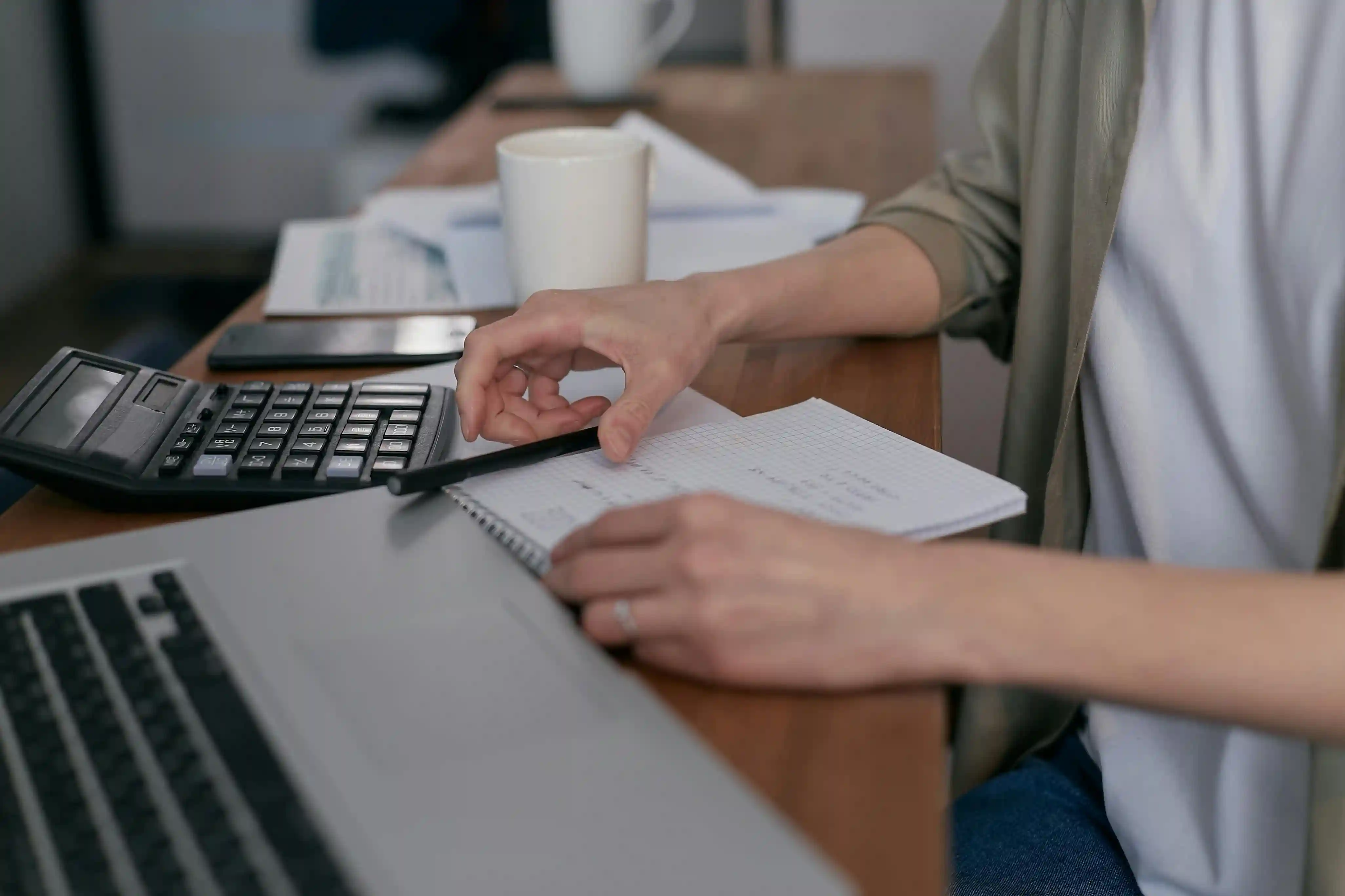 A person working at a desk using a calculator and writing in a notebook beside a laptop and coffee mug.