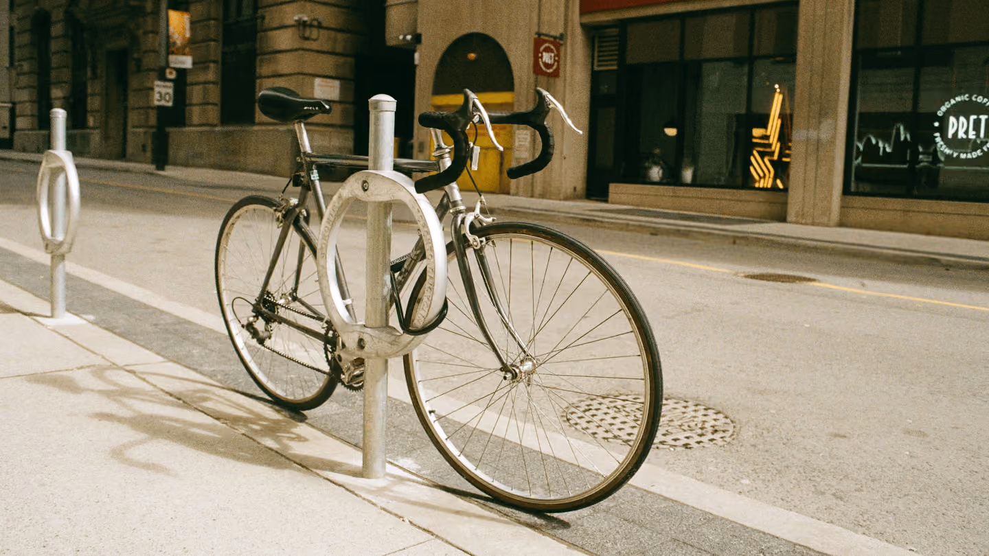 A road bike with drop handlebars locked to a metal ring-style bike rack on an empty city sidewalk.