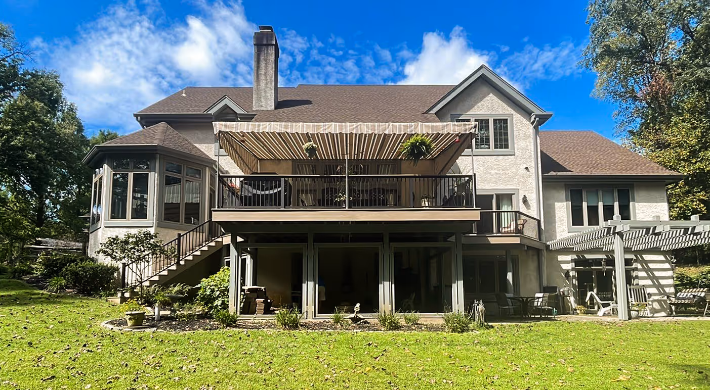 An expansive two-story house with a large deck and a retractable awning, set against a backdrop of lush trees under a clear blue sky.