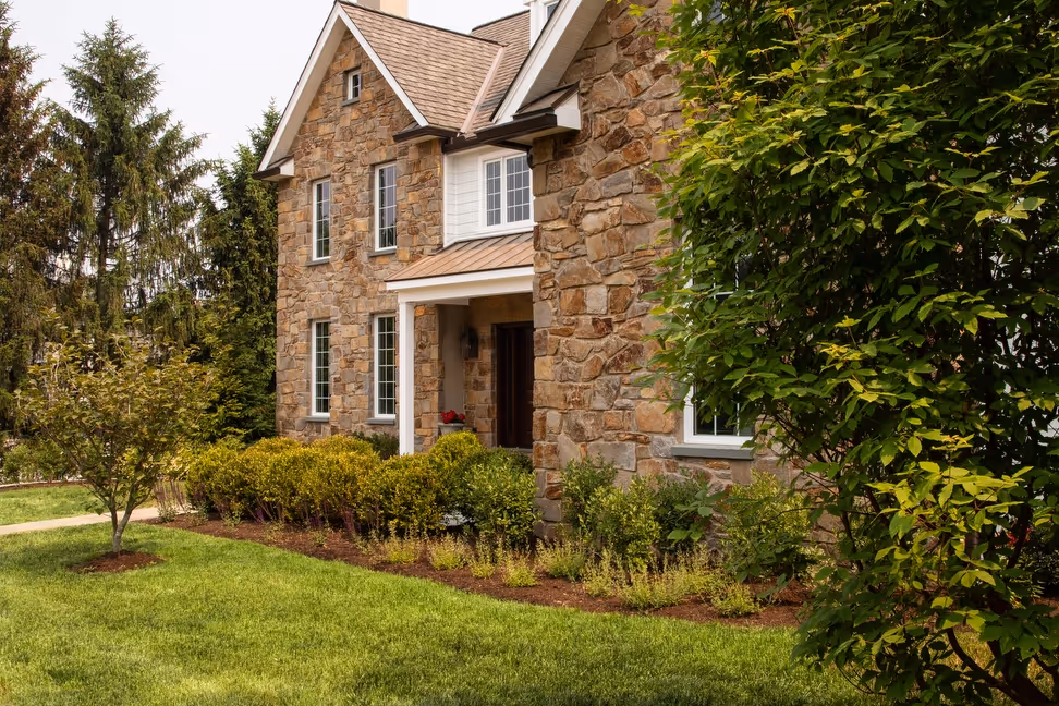 A charming stone-faced house featuring a lush green lawn, decorative shrubs, and vibrant trees, providing a serene and inviting residential atmosphere.