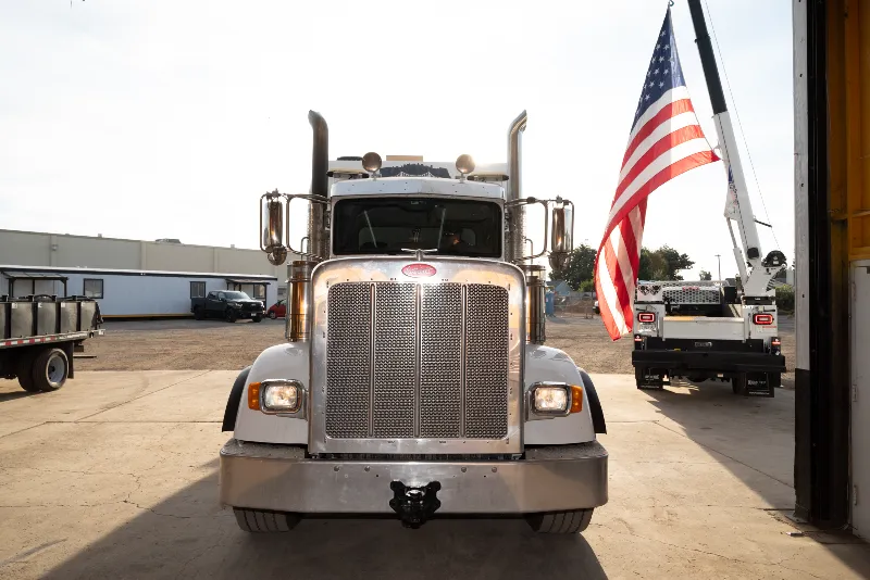 Front-facing Peterbilt semi in service yard, crane truck hoisting US flag behind.