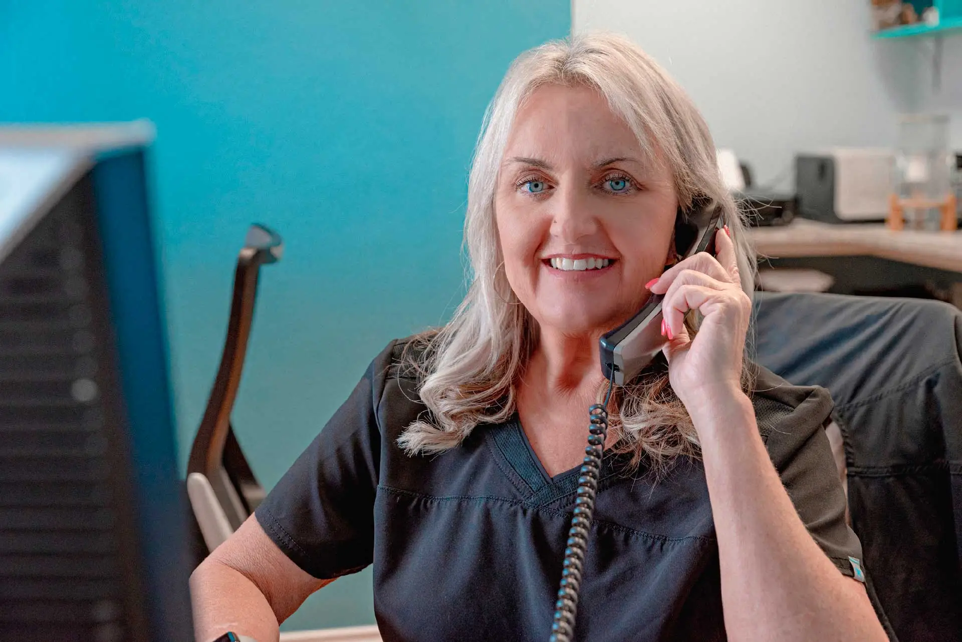Smiling middle-aged woman with blonde hair talking on a corded phone at her desk in an office.