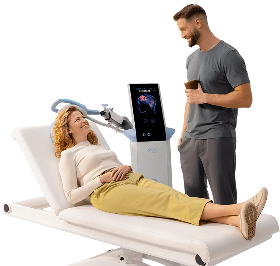Smiling woman lying on a medical examination table with a man standing nearby holding a cup, next to a brain scan machine displaying a brain image.