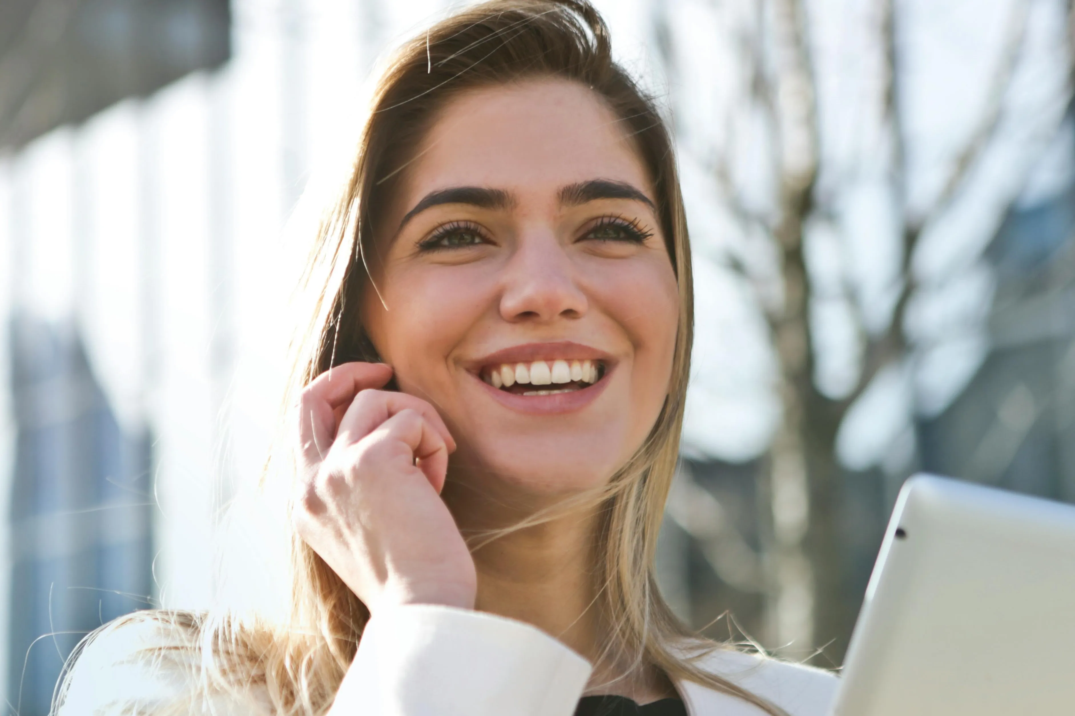 Smiling young woman with blonde hair holding a tablet outside on a sunny day.