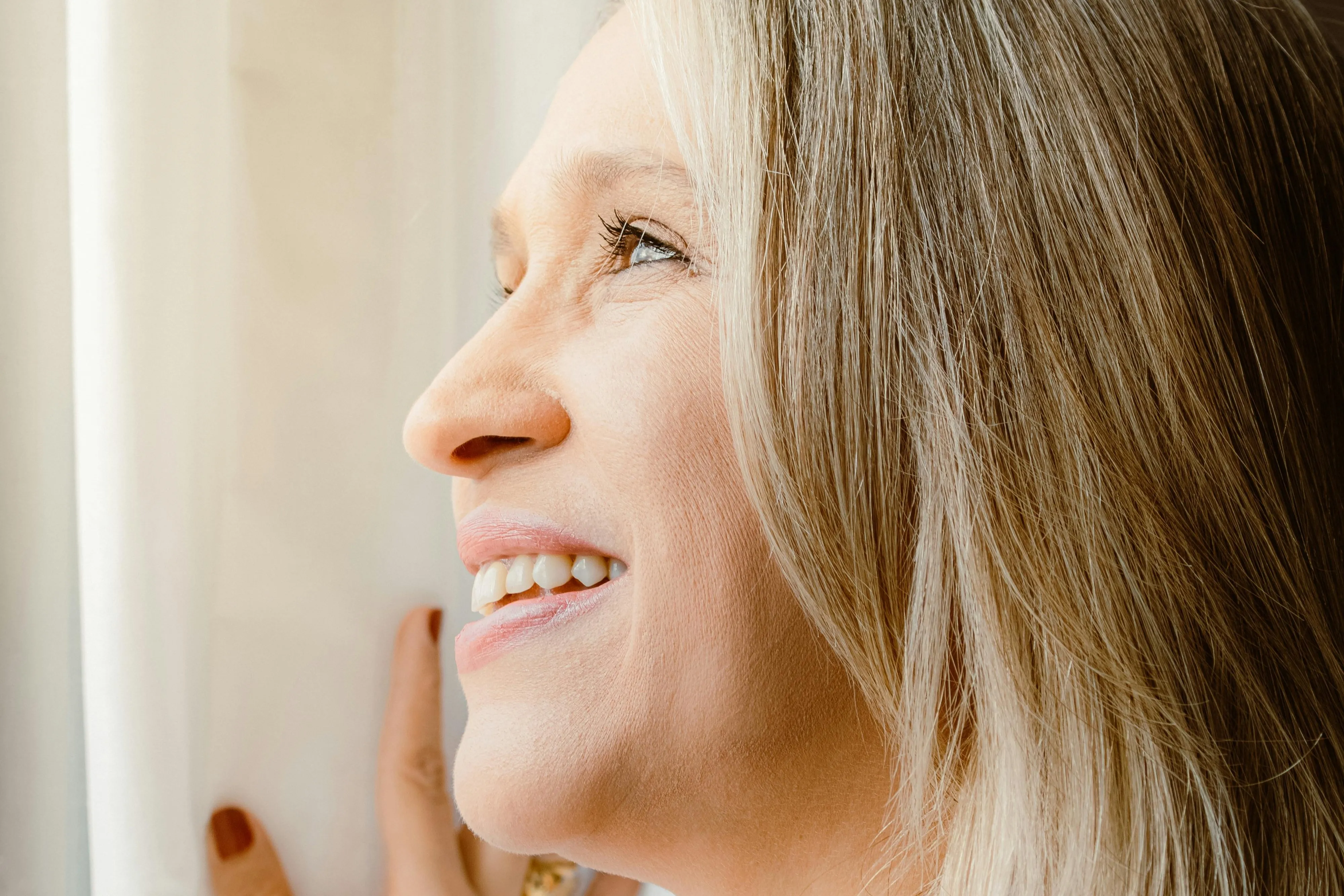 Close-up of a smiling woman with blonde hair looking out a window and touching a white curtain.
