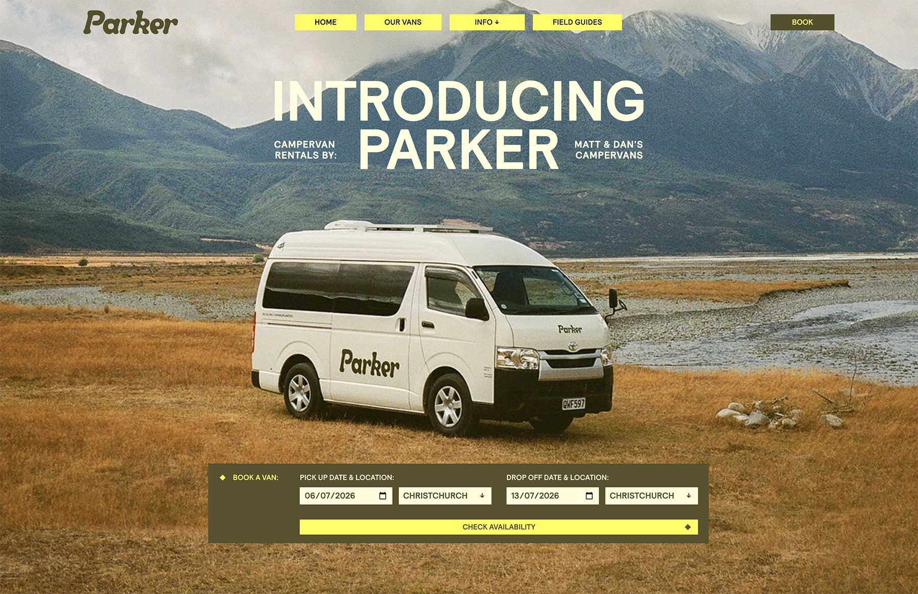 White campervan parked on brown grassland near water with mountains in the background.