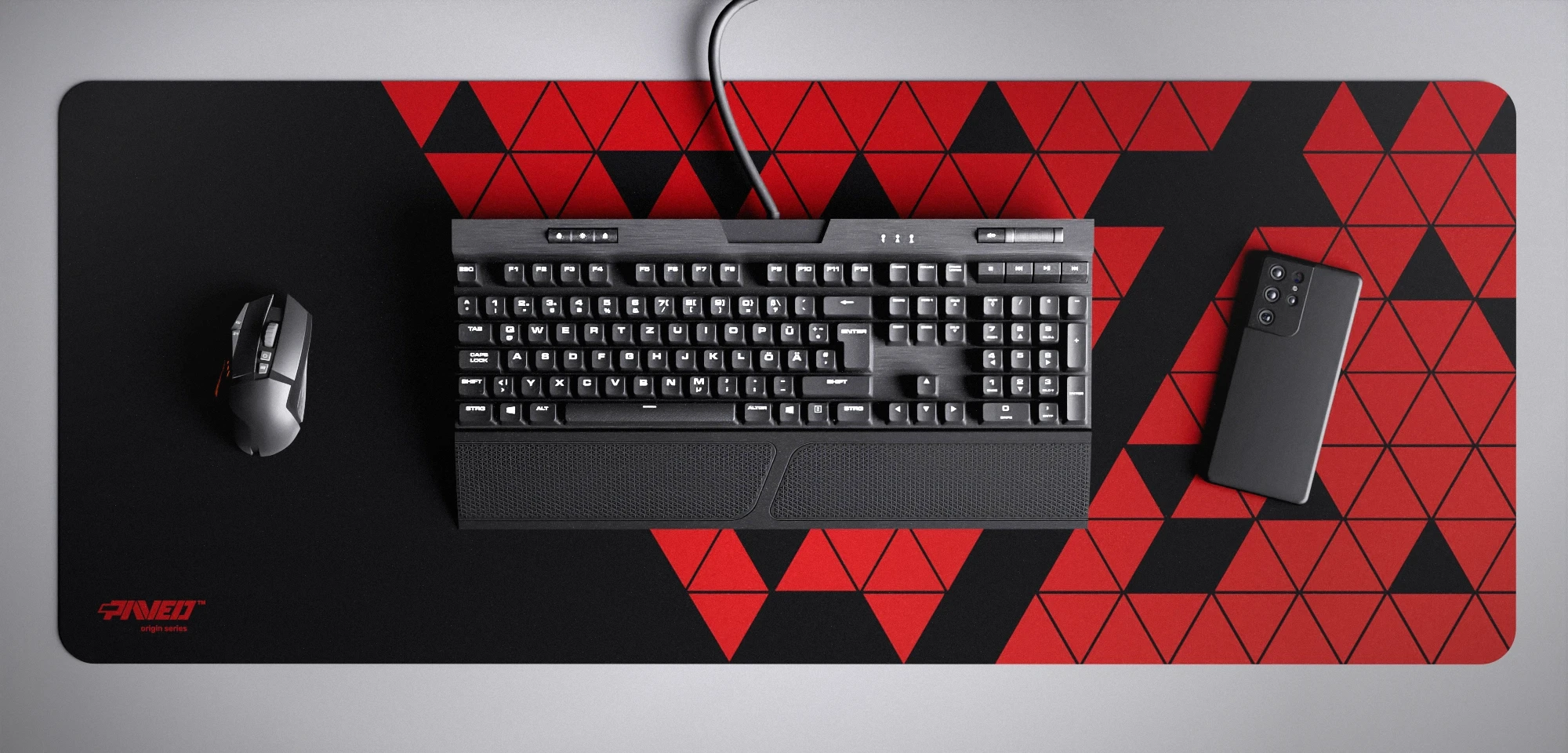 Top view of Night Imperial Prism XXL deskmat 1000mm x 400mm left-handed variant featuring abstract geometric triangular pattern in red and black, water-resistant antimicrobial polyester surface, shown with full-size mechanical keyboard, gaming mouse, and smartphone for scale, ultra-lightweight odour-free AreoFlex microruber base, stitch-free professional finish.