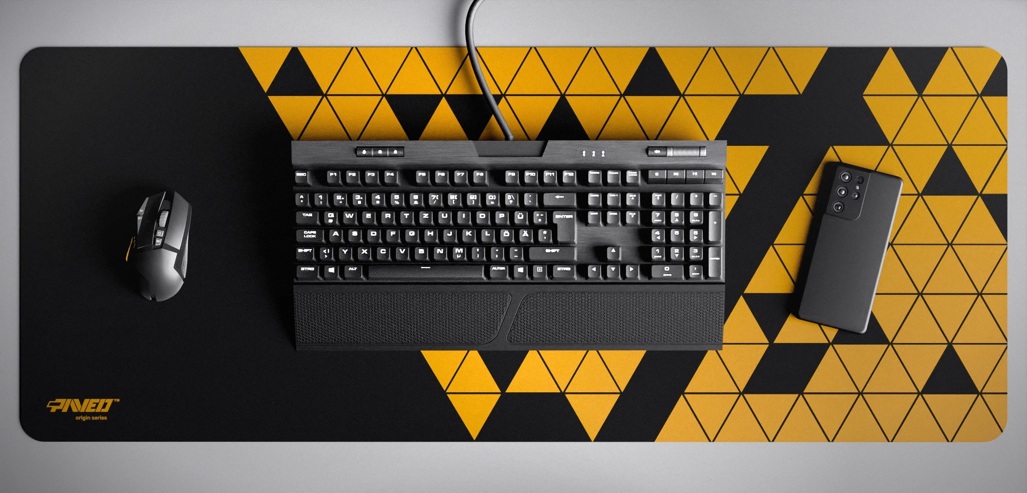 Top view of Night Desert Prism XXL deskmat 1000mm x 400mm left-handed variant featuring abstract geometric triangular pattern in deep yellow and black, water-resistant antimicrobial polyester surface, shown with full-size mechanical keyboard, gaming mouse, and smartphone for scale, ultra-lightweight odour-free AreoFlex microruber base, stitch-free professional finish.