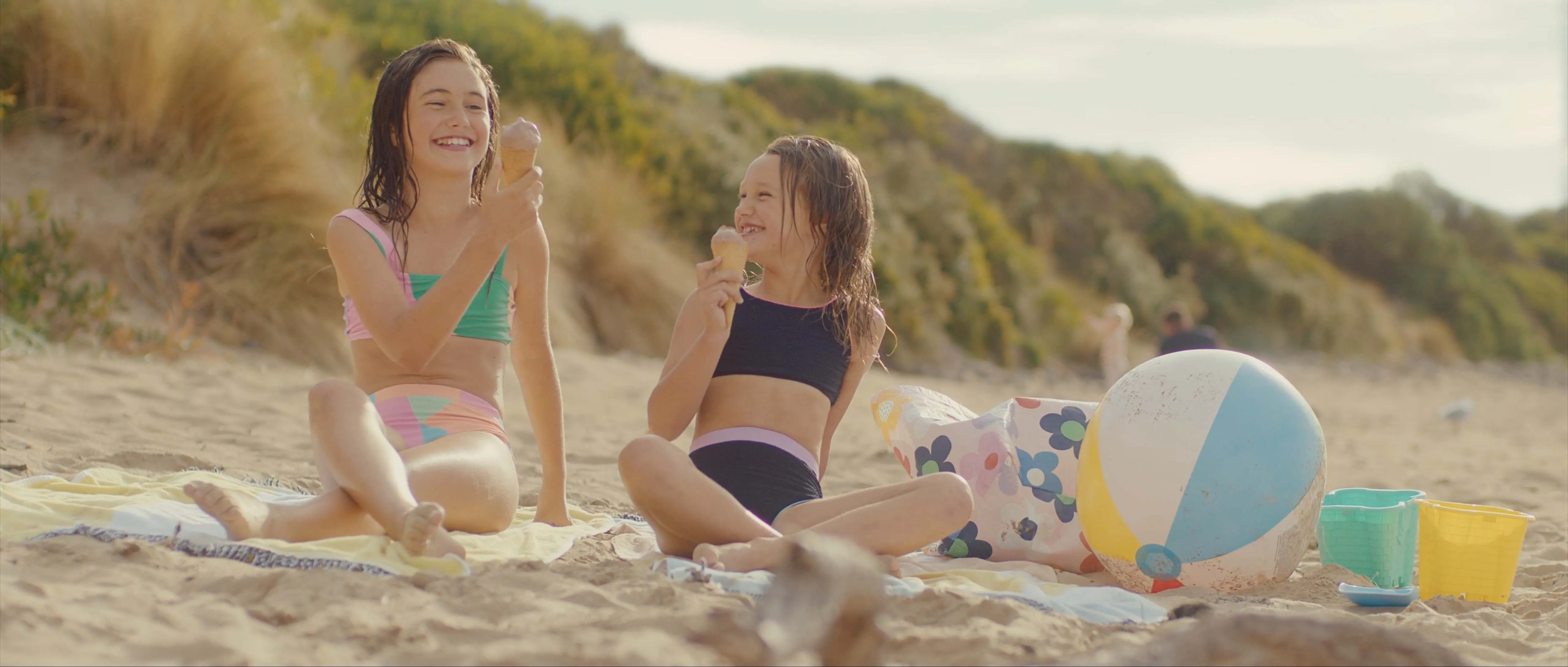 Two kids sitting on the beach eating ice-cream.