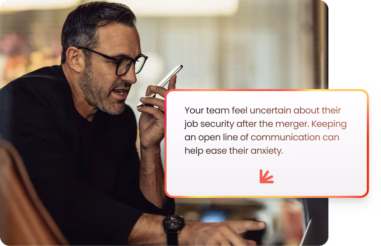 Man with glasses speaking into a smartphone while looking at a laptop, with a message about easing team anxiety by keeping communication open after a merger.