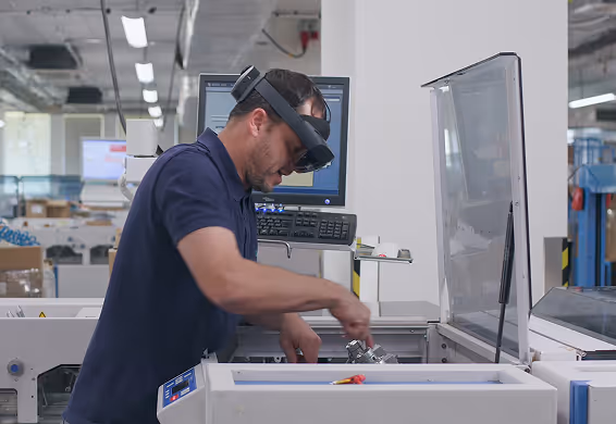 Man in a navy blue shirt wearing augmented reality glasses working on industrial machinery in a factory setting.
