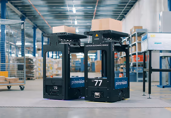 Two black automated warehouse robots carrying cardboard boxes in a spacious industrial storage facility.
