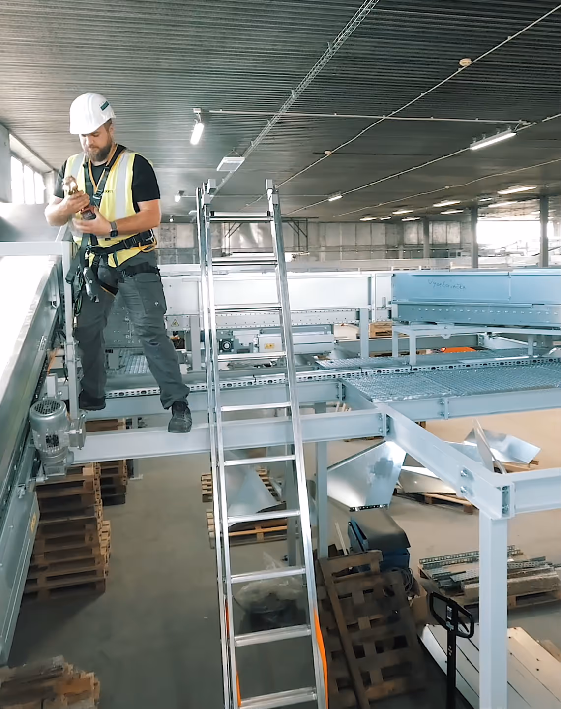 Construction worker in a hard hat and safety harness standing on a steel beam inside an industrial warehouse.