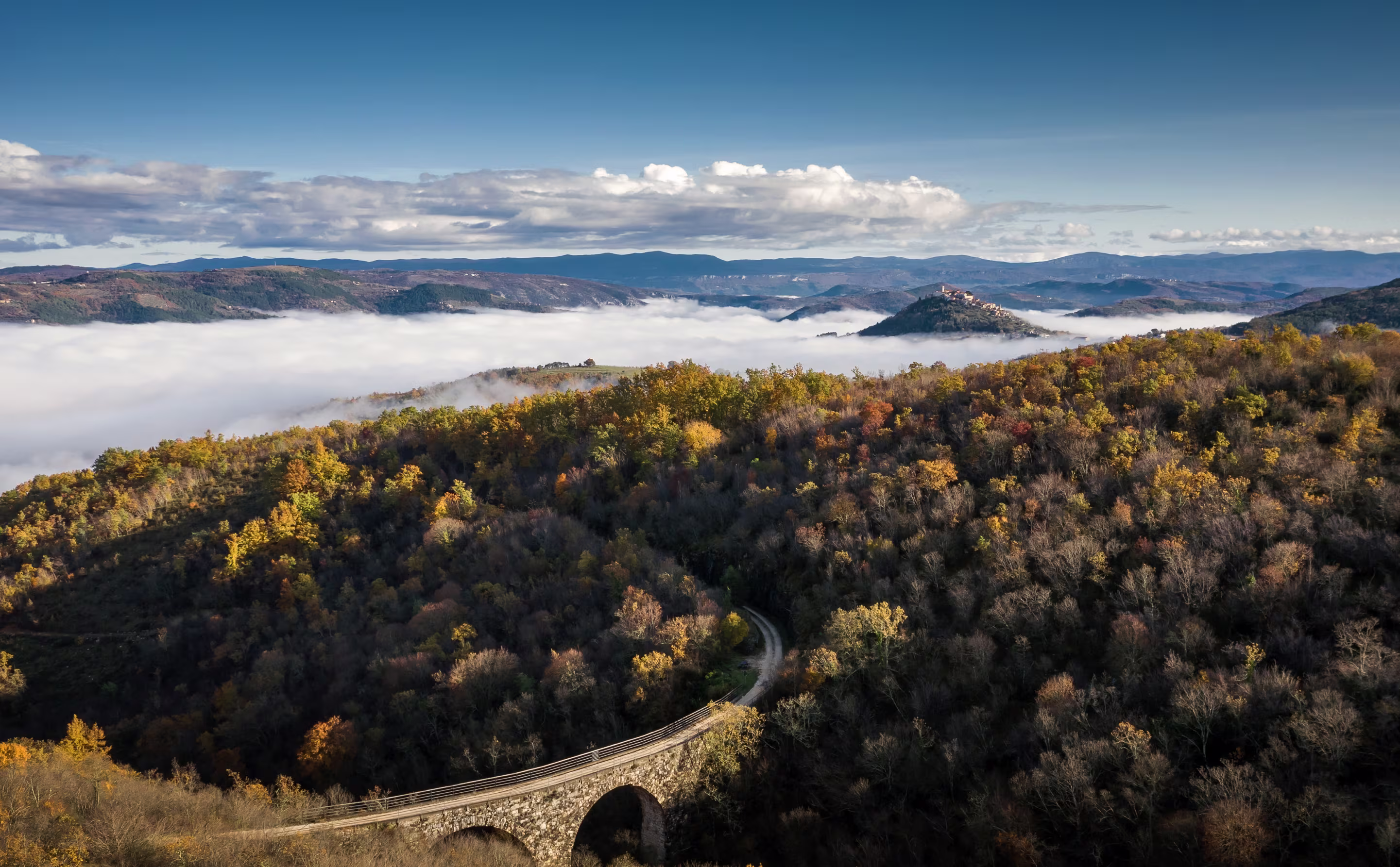 Stone arch bridge over forested hills with autumn foliage under a blue sky and mist in the valley.