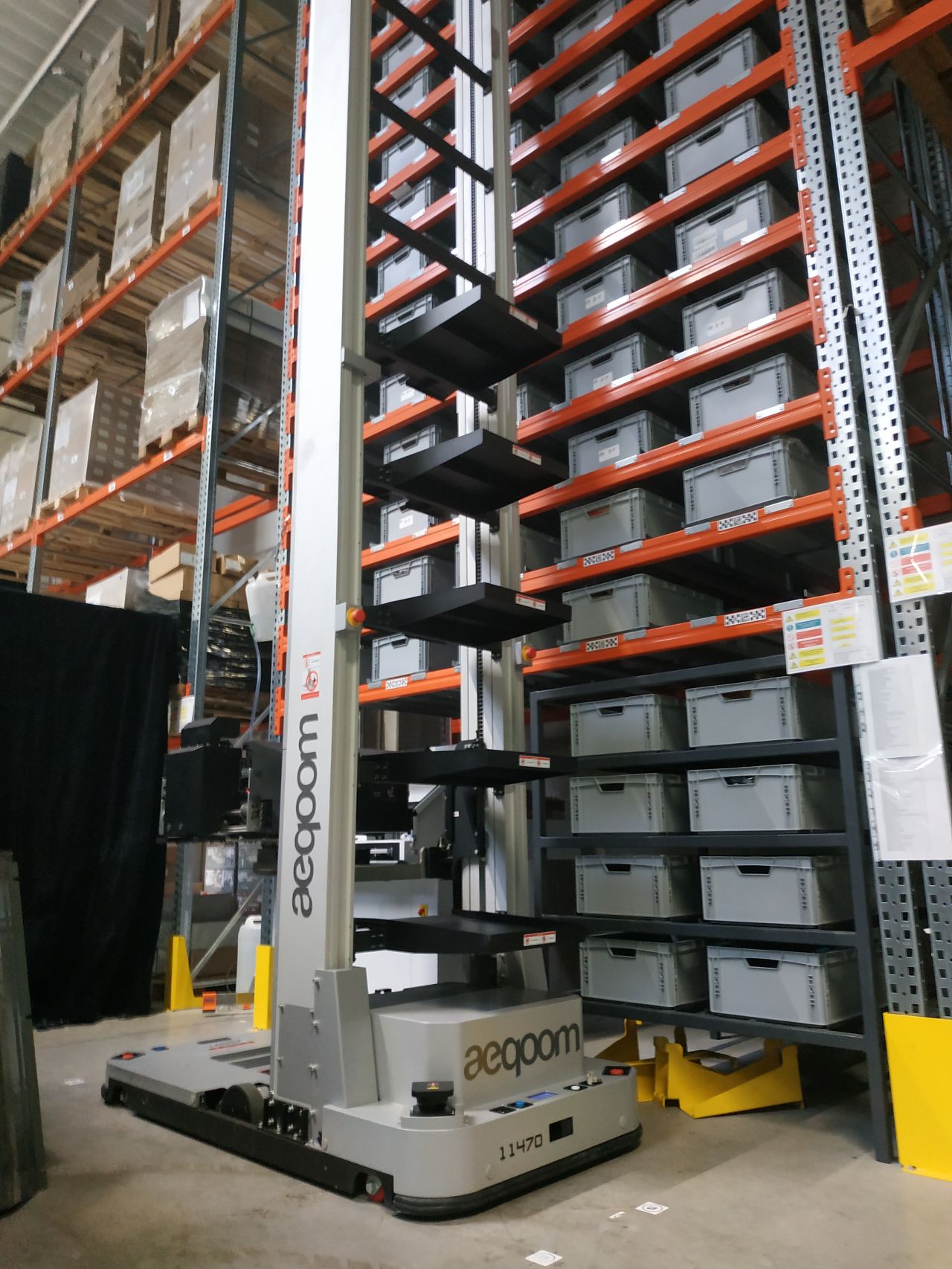 Automated storage and retrieval robot labeled 'aecqom' operating in a warehouse aisle with stacked gray storage bins on orange shelves.