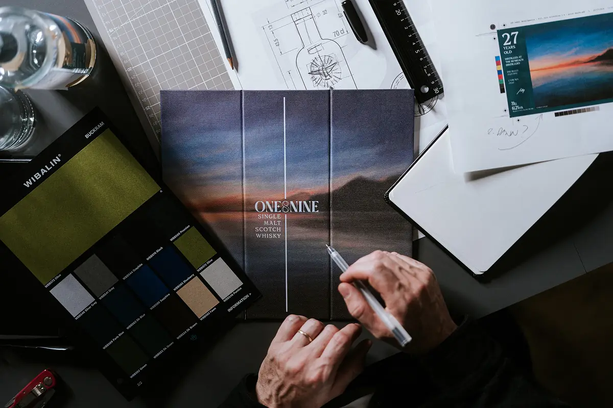 Designer Tom working on the packaging concept for One8Nine’s The Eternal Cycle whisky, reviewing material swatches, label artwork, and bottle design sketches on his desk.
