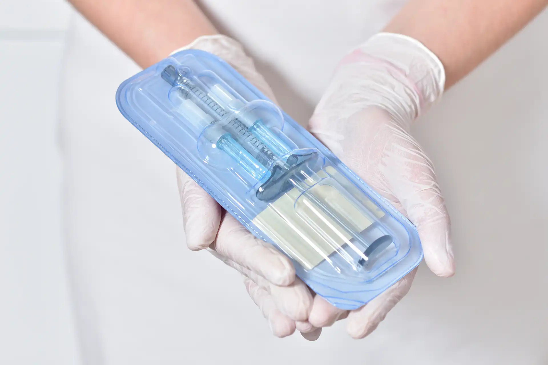 Gloved hands holding a sealed plastic package containing a syringe medical device.