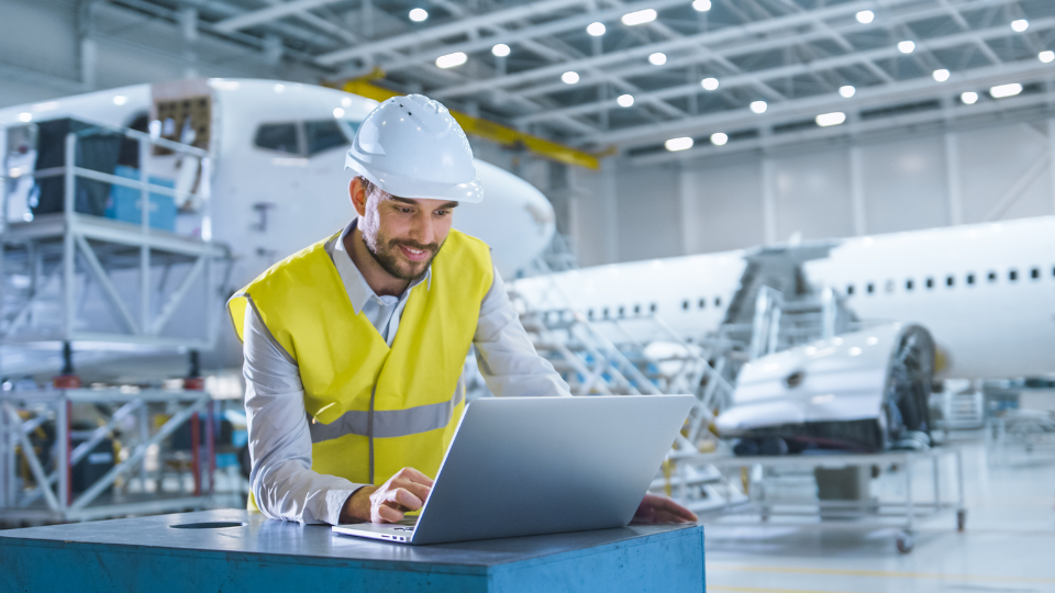 An aviation professional in safety gear using a laptop in an aircraft maintenance facility.