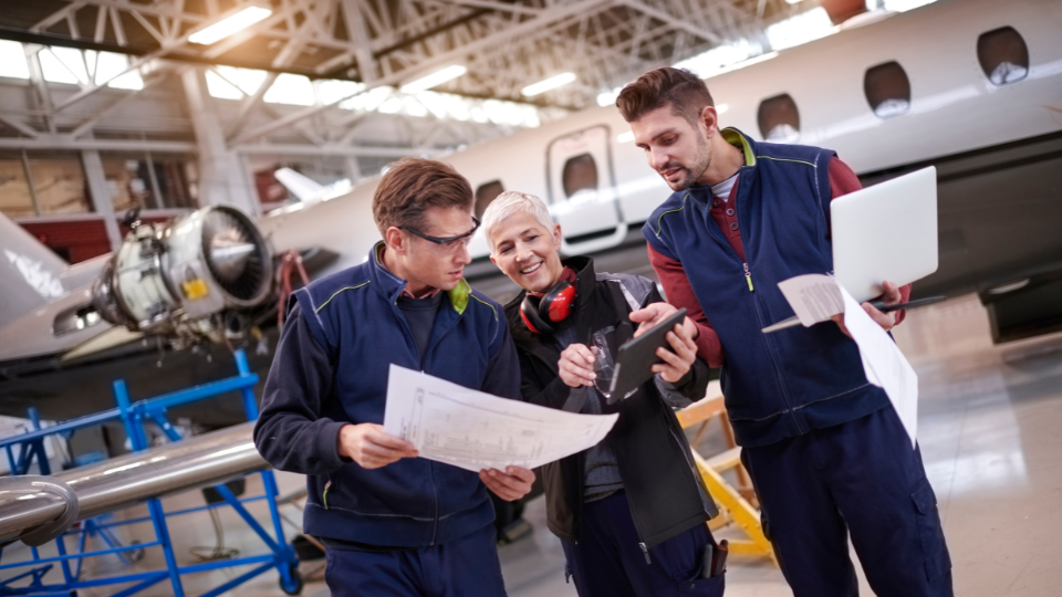 A team of aviation professionals, including maintenance staff and operational coordinators, reviewing software and documentation in an aircraft maintenance hangar.