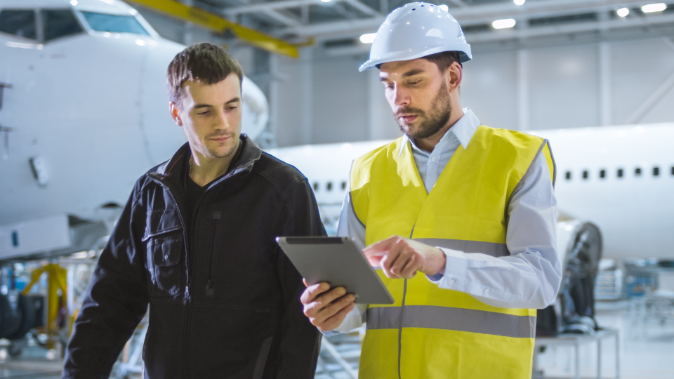 Two aviation professionals are looking at a digital tablet while discussing operational tasks in an aircraft maintenance environment.