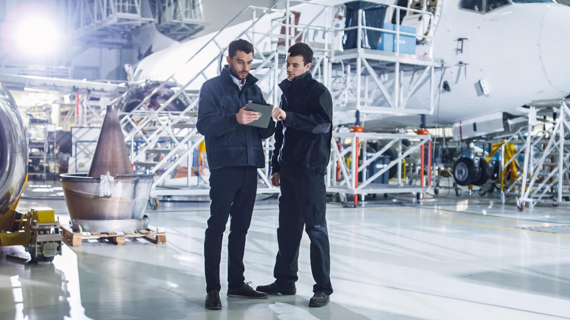 Two aircraft maintenance engineers in a hangar discussing information on a tablet, with an aircraft undergoing detailed inspection and servicing in the background.
