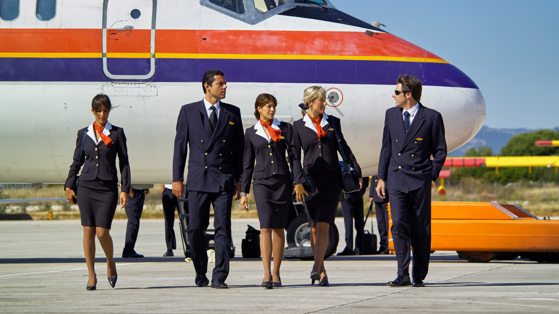 A flight crew, including pilots and cabin crew, walking together on the tarmac in uniform beside a passenger aircraft.