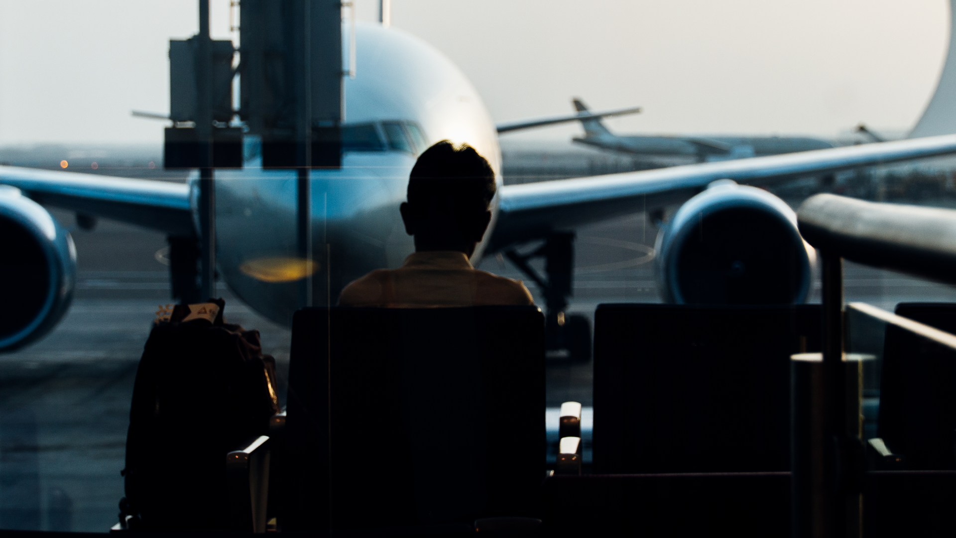 A person is seated in an airport waiting area, looking out a large window at a passenger airplane parked on the tarmac.