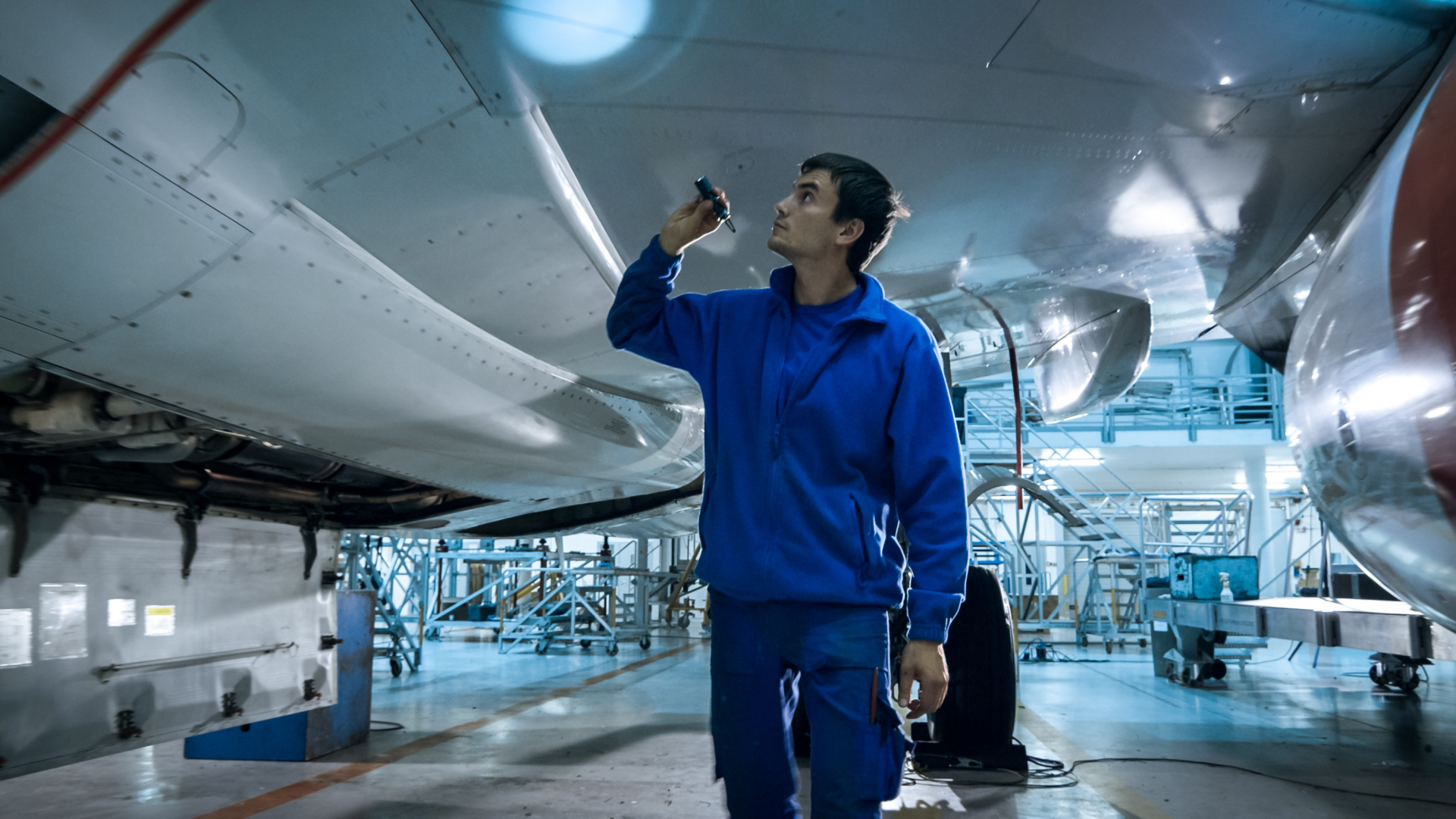 An aircraft mechanic in a blue uniform stands under the wing of an airplane in a hangar, holding a flashlight to inspect the aircraft's structure.