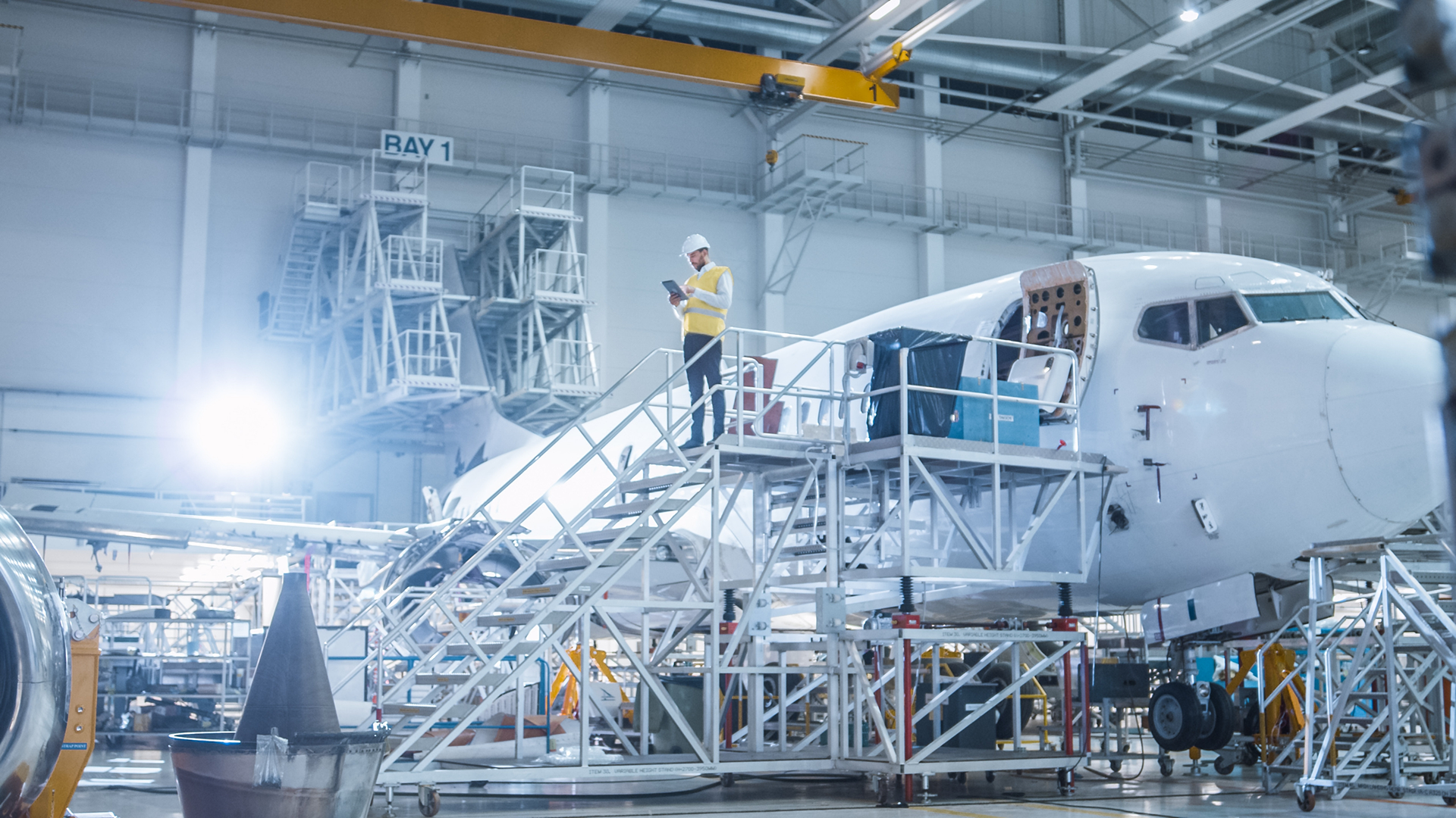 An aircraft maintenance worker stands on scaffolding next to a large airplane in a hangar, looking at a tablet.