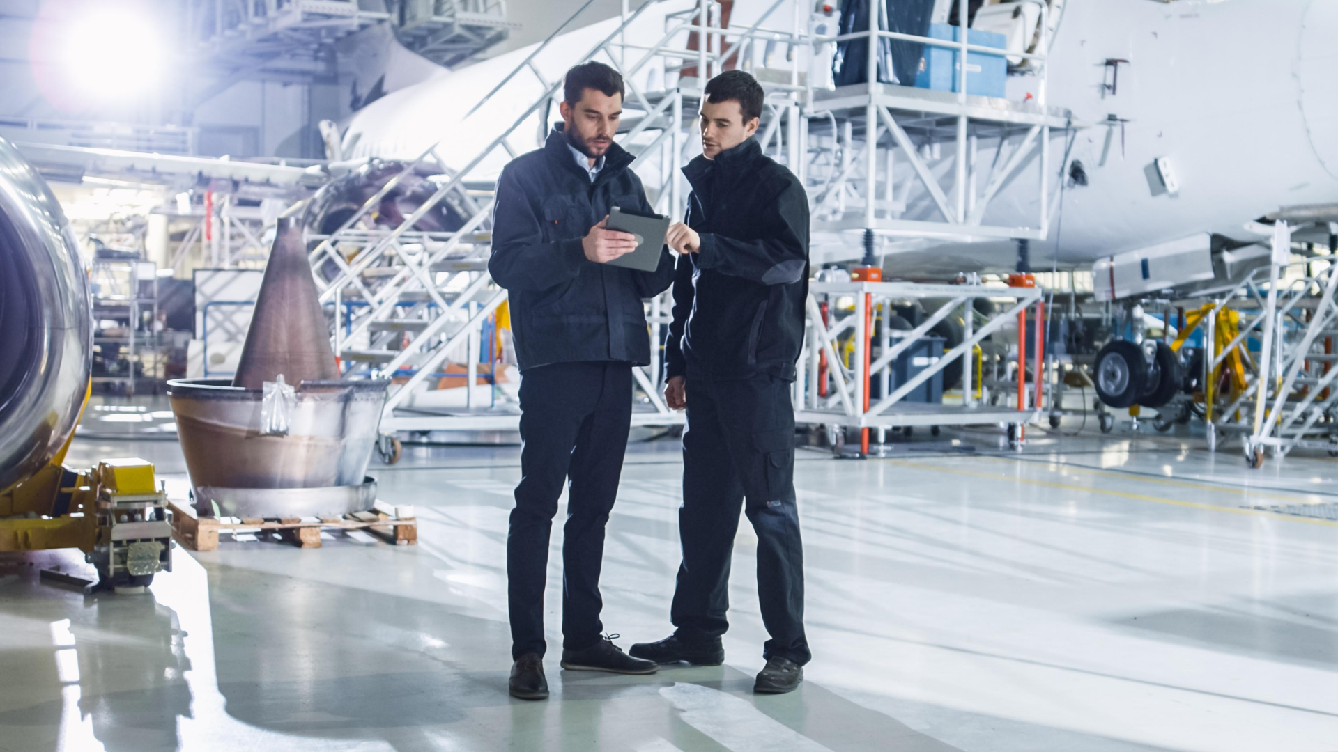 Two aircraft maintenance technicians stand in a large hangar next to an airplane, discussing information on a digital tablet.