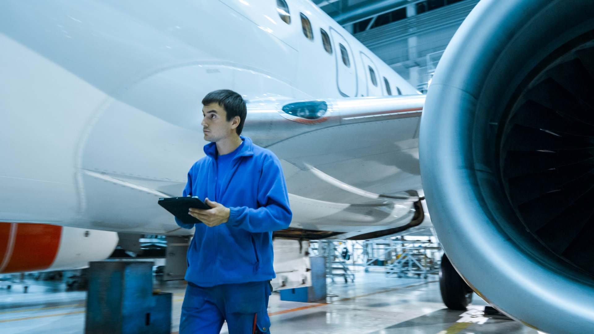 A maintenance technician stands near an airplane engine and fuselage in a hangar, holding a tablet.