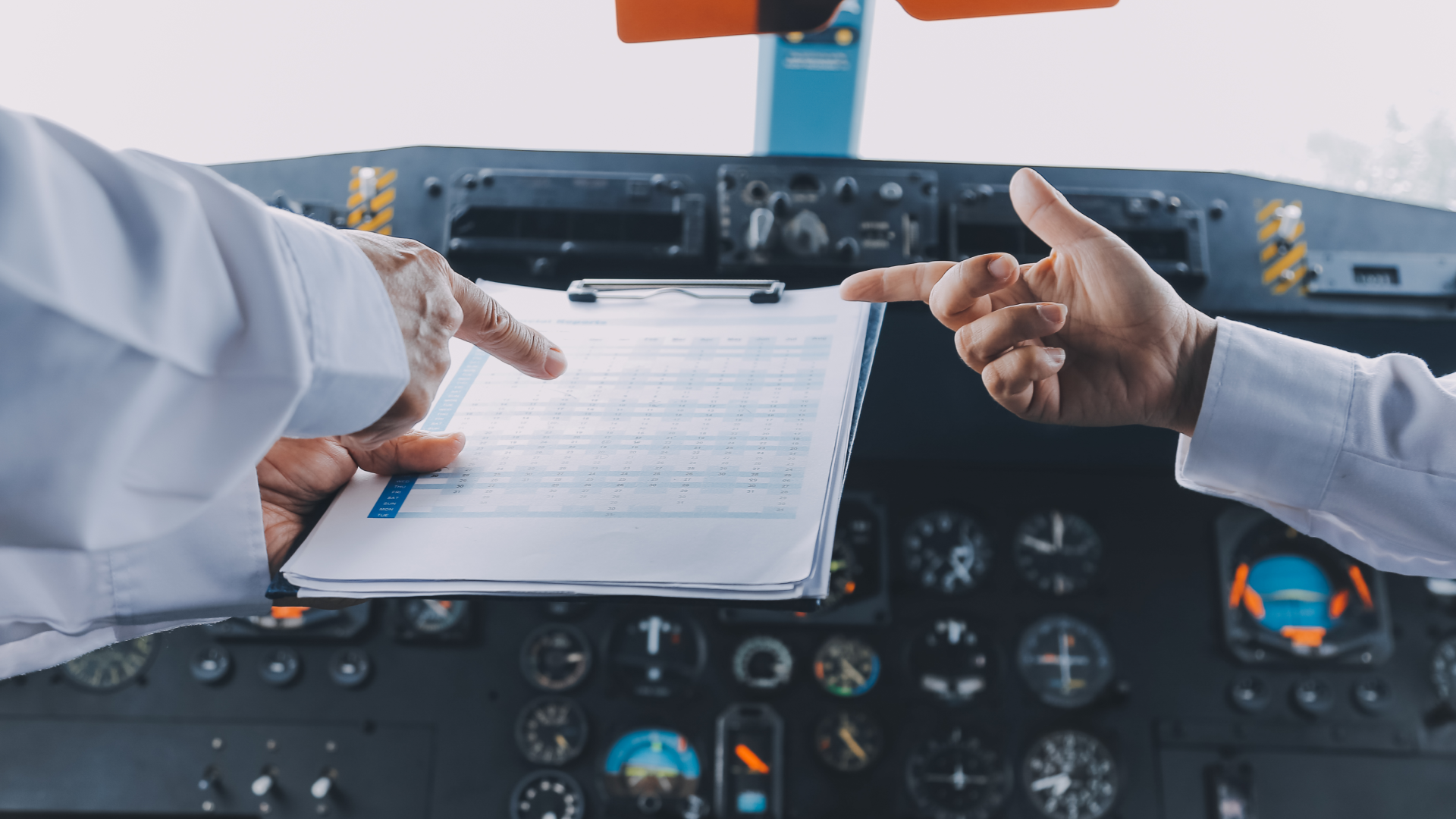 Two people point at a spreadsheet on a clipboard while in an aircraft cockpit.
