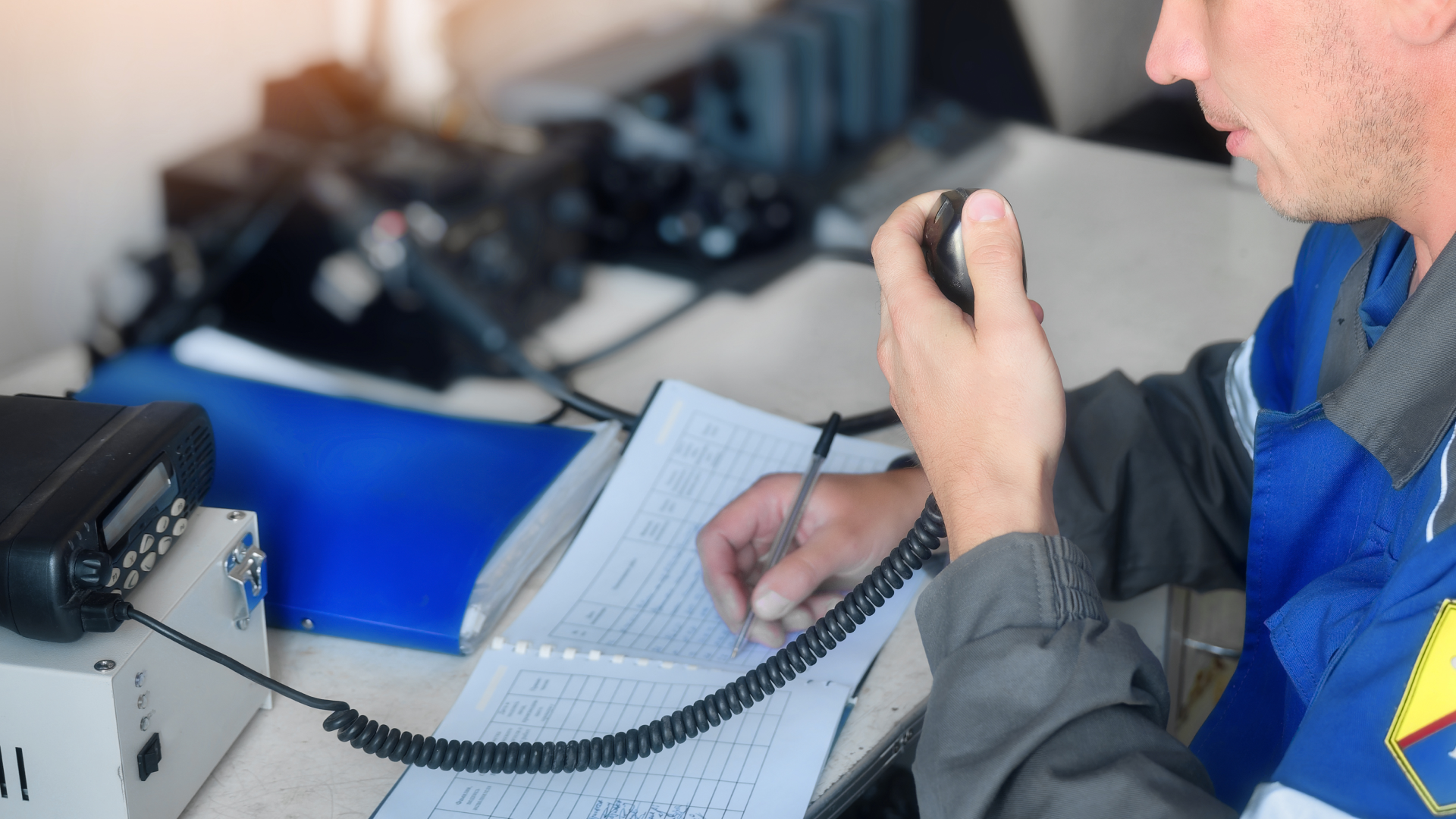 A person in a work uniform speaking into a handheld radio while writing notes in a logbook at a desk.